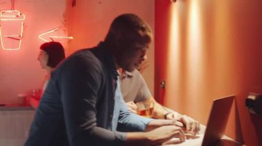 Orta doğulu erkek arkadaşı çay fincanı alıp ona doğru yürürken ve projeyi tartışırken, genç Afrikalı Amerikalı bir adam modern çatı katında bilgisayar üzerinde çalışıyor.