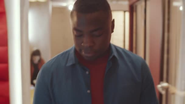 身穿休闲装的年轻非洲裔美国人走在合作办公空间的走廊上 一边用智能手机 一边打招呼同事 — 图库视频影像