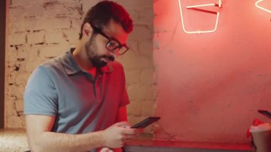 Orta Doğulu genç bir adamın gözlüklü ve günlük kıyafetleriyle akıllı telefondan yazı yazarken ve çatı katı duvarının yanında neon ışıkla dururken kameraya bakarken belini kaldır.