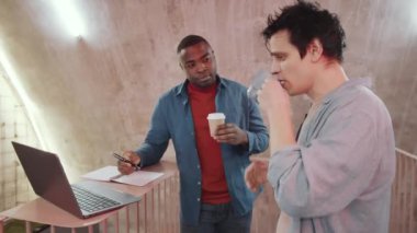Kafkasyalı ve Afrikalı genç erkek meslektaşlar kahve ve çay içerek modern çalışma ofisinde çalışırken dizüstü bilgisayarla projeyi tartışıyorlar.