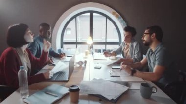 Dizüstü bilgisayar kullanan ve belgeleri paylaşan çok ırklı genç meslektaşlardan oluşan yaratıcı bir ekip, birlikte çalışma odasının penceresinin yanındaki masada proje üzerinde çalışırken,