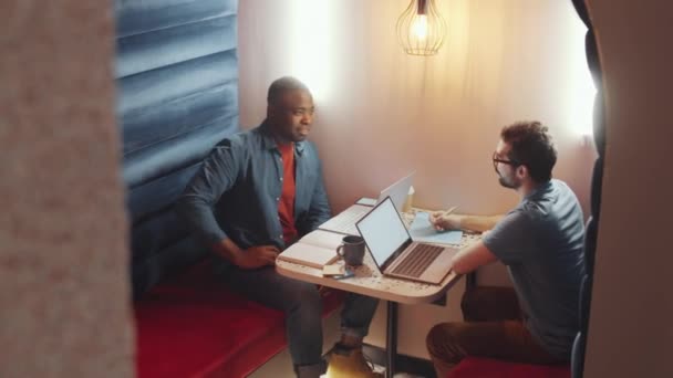 男性同僚とノートパッドでノートを議論しながら カフェからリモートで働く若いアフリカ系アメリカ人男性の高角度ショット — ストック動画