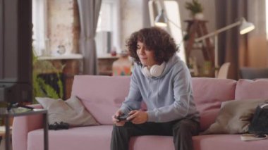 Karışık ırk genci evdeki kanepede oturuyor, gülümsüyor ve konsolda video oyunu oynuyor.