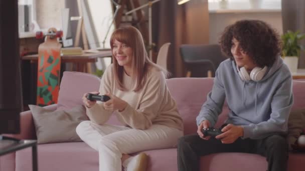 美しいです女性と彼女の十代の息子笑顔と使用していますコントローラーゲームをプレイ中にソファに家 — ストック動画