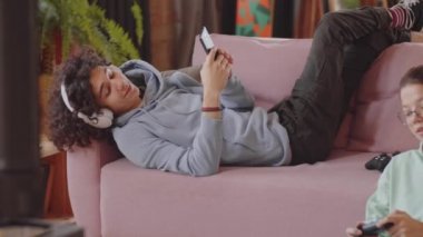 Genç çocuk kanepede uzanıyor, akıllı telefon kullanıyor ve kablosuz kulaklıkla müzik dinliyor. Bu sırada kız kardeşi yerde oturuyor ve evde konsol oyunu oynuyor.