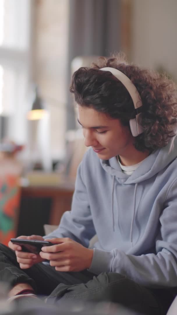 自宅でソファに座っている無線ヘッドフォンでハンサムな十代の男の子の垂直形式のビデオ 笑顔とスマートフォンでモバイルゲームをプレイ — ストック動画