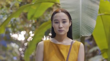 Çekici, esmer, genç Asyalı bir kadının serada tropik yeşil bitkilerin arasında dikilirken kameraya özgüvenle baktığı göğüs üstü fotoğraf.