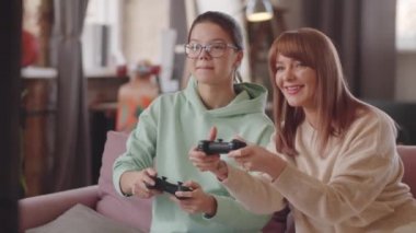Evde oturup gülümseyen ve sohbet eden anne ve genç kız konsolda oyun oynuyorlar.