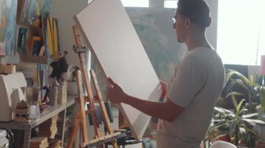 Erkek sanatçı resim sehpasına doğru yürür ve yaratıcı stüdyoda çalışmaya hazırlanırken boş tuval yerleştirir.