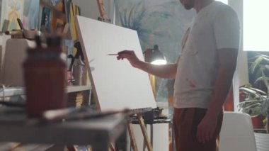 Resim stüdyosunda duran ve kurşun kalemle tuvale resim çizen lekeli tişörtlü yaratıcı genç adamın görüntüsünü yakınlaştır.