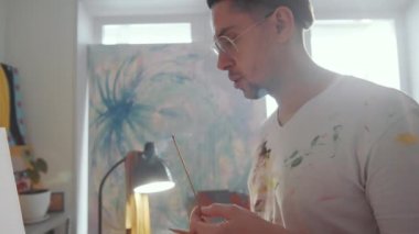 Lekeli elbiseli bir adam tütsü çubuğunun ucunu yakıyor, alevi söndürüyor ve tutacağına koyuyor, sonra da resim stüdyosunda resim çiziyor.