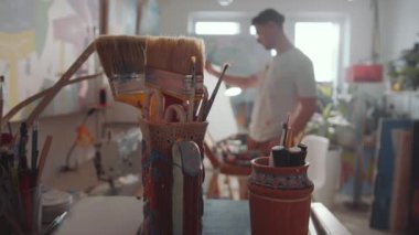 Sanat atölyesinde el yapımı vazoların resim fırçaları, kalemler ve çizim araçları ile yakın plan çekimini yapın; erkek sanatçı odaklı olmayan arka planda resim üzerinde çalışıyor