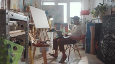 Yaratıcı stüdyoda sandalyede oturan ve dizüstü bilgisayarda yazan lekeli elbiseli genç bir erkek sanatçının fotoğrafını yakınlaştır.