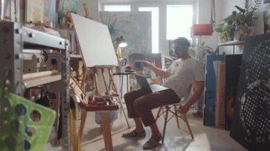 Genç erkek sanatçının yaratıcı stüdyoda oturduğu, el salladığı, konuştuğu ve resim gösterdiği görüntüyü yakınlaştırın.