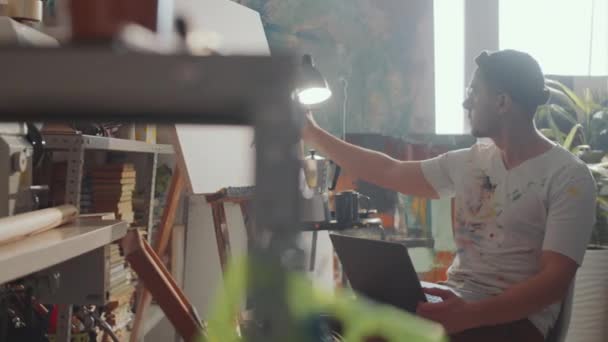創造的なスタジオに座っているプロの男性アーティスト 彼の膝の上にラップトップを保持し キャンバス上の鉛筆で描く — ストック動画