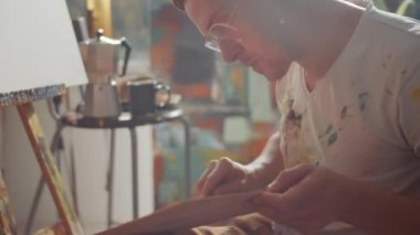 Profesyonel erkek ressamın ahşap palet tutarken ve palet bıçağıyla boya renklerini karıştırırken yaratıcı stüdyo çalışmalarına hazırlanırken elde çekilen kamera görüntüsü.