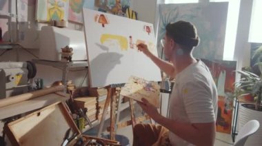 Yaratıcı erkek sanatçının ahşap paleti tutarken ve palet bıçağıyla tuvale boya sürerken stüdyoda yeni bir resim çizerken görüntüsünü yakınlaştırın.