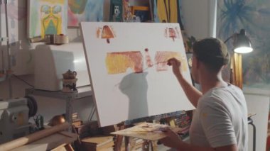 Stüdyoda oturan, elinde ahşap palet olan ve fırçayla tuvale resim çizen erkek ressamın arka planı