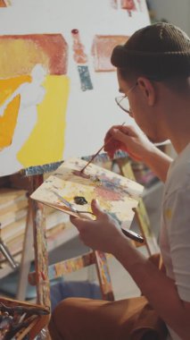 Erkek ressamın tahta palet taşıdığı ve stüdyoda tuvale fırçayla resim yaptığı omuz üstünden dikey çekim.