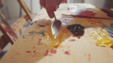 Profesyonel ressamın elleri yakın çekim. Sarı boyayla palet bıçağını ahşap palette karıştırıyor.