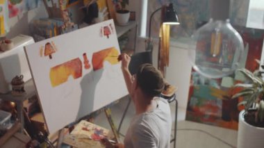 Şapkalı yaratıcı bir adamın ahşap palet tutarken ve sanat stüdyosunda tuvale fırçayla resim yaparken yüksek açılı çekimi.