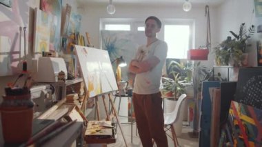 Resim stüdyosunda kolları çapraz duran ve kameraya poz veren lekeli tişörtlü genç bir erkek sanatçının görüntüsünü yakınlaştır.