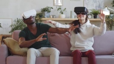 Genç Afro-Amerikalı adam ve VR kulaklıklı melez kadın oturma odasında kanepede oturuyor, gülümsüyor ve bir şeylere dokunurken artırılmış gerçekliğin tadını çıkarıyorlar.