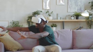VR kulaklıklı genç, gülümseyen Afro-Amerikalı adam evdeki kanepede oturuyor ve sanal gerçekliği aletle yaşarken havada bir şeylere dokunuyor.