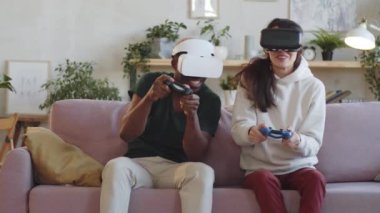 VR kulaklıklı neşeli genç bir çift evdeki kanepede oturmuş oyun kağıtları kullanıyor ve heyecanla video oyunu oynuyorlar.