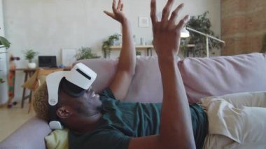 VR kulaklıklı neşeli siyahi adamın oturma odasında kanepede uzanmış, gülümseyen ve havaya dokunurken görüntüsünü aşağı eğ.