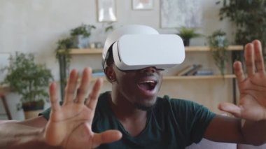 Genç Afro-Amerikalı adam VR kulaklığını takıyor, ellerini uzatıyor, evde video oyunu oynarken heyecanla etrafa bakıyor ve gülümsüyor.