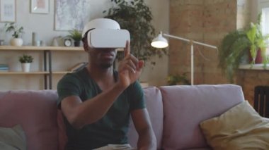 VR kulaklıklı genç Afro-Amerikalı adam evdeki koltukta oturuyor, gelişmiş gerçeklikteki etkileşimli arayüzü kullanarak görünmez dokunmatik ekrana dokunuyor ve ona dokunuyor.