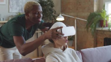 Neşeli siyahi bir adam karısının kafasına VR kulaklığı takıyor ve evde alet kullanırken onun artırılmış gerçeklik hakkındaki heyecanını izlerken gülümsüyor.