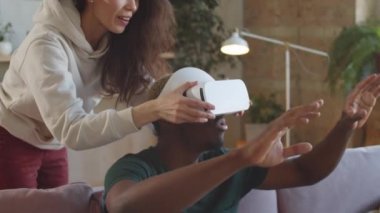 Genç melez kadın, Afro-Amerikan bir adamın kafasına VR kulaklığı takıp gülümseyerek evdeki artırılmış gerçeklik için heyecanlanmasını seyrediyor.