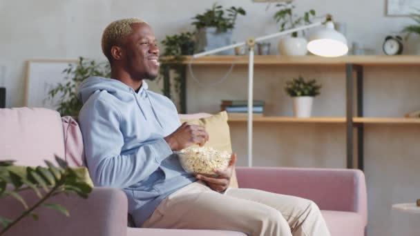 若い幸せなアフリカ系アメリカ人の男はソファに座って ポップコーンを食べ 家でテレビで映画を見ながら笑って — ストック動画