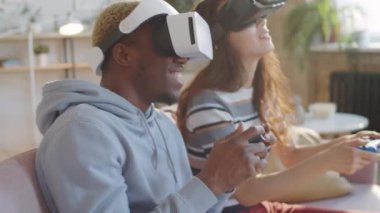 VR kulaklıklı genç ve çok ırklı bir çift koltukta oturmuş, oyun kağıtları kullanıyor ve evde video oyunu oynuyorlar.