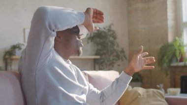 VR kulaklıklı neşeli Afro-Amerikalı genç adam evde alet kullanırken gülümsüyor ve havaya dokunuyor.