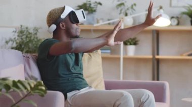 Genç Afrikalı Amerikalı adam evdeki koltukta oturmuş video oyunu oynarken havada bir şeylere dokunuyor.