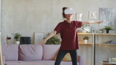 Spor kıyafetli genç bir kadın. Oturma odasında gülümsüyor ve egzersiz yapıyor. Evde VR kulaklıkla egzersiz yapıyor.