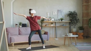 VR kulaklıklı genç bir kadın evde paspas üzerinde ileri doğru pratik yaparken artırılmış gerçeklikte fitness uygulaması kullanıyor.