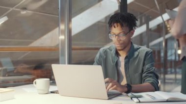 Gündelik giyinmiş genç Afrikalı Amerikalı bir adam dizüstü bilgisayar kullanıyor ve sonra kameraya bakıp gülümsüyor. Şirket ofisinde cam duvarlarla çalışırken.