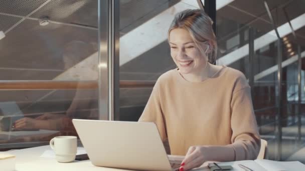 坐在现代办公室的办公桌前 戴着无线耳机的金发美女一边微笑着 一边用笔记本电脑通过视频通话聊天 — 图库视频影像