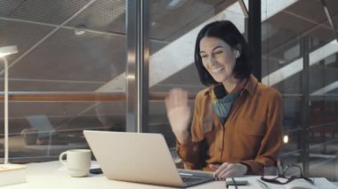 Pozitif esmer iş kadını, modern ofiste çalışırken kablosuz kulaklık takıyor, gülümsüyor, el sallıyor ve video aracılığıyla konuşuyor.