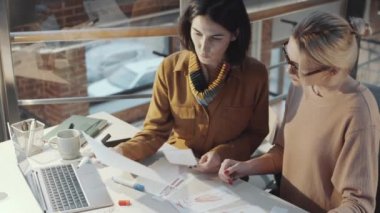 Yüksek açılı esmer ve sarışın terzilerin stüdyoda masasında oturup kıyafet tasarımı çizimlerini tartışmaları.