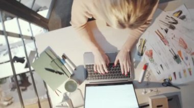 Dizüstü bilgisayarda yazan kadın moda tasarımcısının üst görüntüsü ve stüdyoda masa başında çalışırken keçeli kalemle yeni kıyafet koleksiyonunun çizimleri.