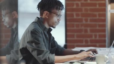 Genç Afro-Amerikan bir adamın gözlüklü ve günlük kıyafetleri içinde dizüstü bilgisayar kullanırken, kameraya bakarken ve çatı katında cam duvarlı bir masada çalışırken gülümserken.