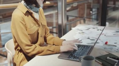 Modern stüdyoda çizimler, fincanlar ve not defterleriyle masa başında çalışırken dizüstü bilgisayarda yazan kadın moda tasarımcısının orta bölüm fotoğrafı.