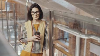 Gecelikli ve bardaklı mutlu esmer iş kadını kahve bardağını tutuyor, kameraya bakıyor ve ofiste cam duvara yaslanırken gülümsüyor.