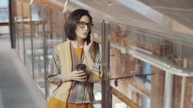 İş yerinde cam duvarın yanında dikilirken kahve fincanı tutan, gülümseyen ve telefonla konuşan günlük giysili, gözlüklü beyaz bir iş kadınının yüksek açılı çekimi.