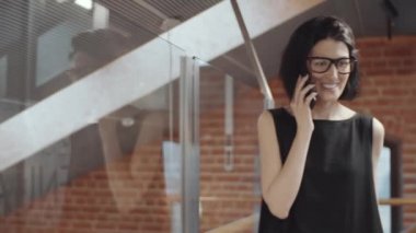 Modern çatı katında cam duvar boyunca yürürken siyah elbiseli, gözlüklü, gülümseyen ve cep telefonuyla konuşan esmer iş kadınının belden yukarısını çek.